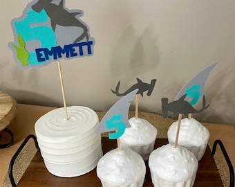 May include: A white cake with a blue and green shark topper that says "Emmett" on it. There are four cupcakes with shark fin toppers and a number "5" on them. The cupcakes and cake are on a wooden tray.