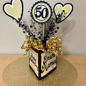 May include: A gold and black anniversary centerpiece with the number 50, hearts, and star accents. The centerpiece has the words "Happy Anniversary" and "Insert Names" on the sides. The centerpiece is on a gold placemat.