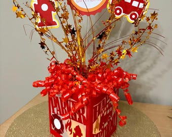 Op de afbeelding: Een rood en geel verjaardagsmiddelpunt met een brandweerthema. Het middelpunt heeft een rode doos met uitgesneden ontwerpen, waaronder een brandkraan en de tekst "Happy Birthday". Het is versierd met stervormige accenten en een "Happy Birthday"-bord.