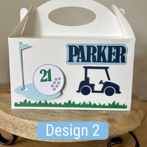 Puede incluir: Caja de regalo blanca con tem&aacute;tica de golf. La caja tiene una bola de golf verde y azul con el n&uacute;mero 21, una bandera de golf verde, un carrito de golf azul y el nombre "Parker" en la parte delantera.