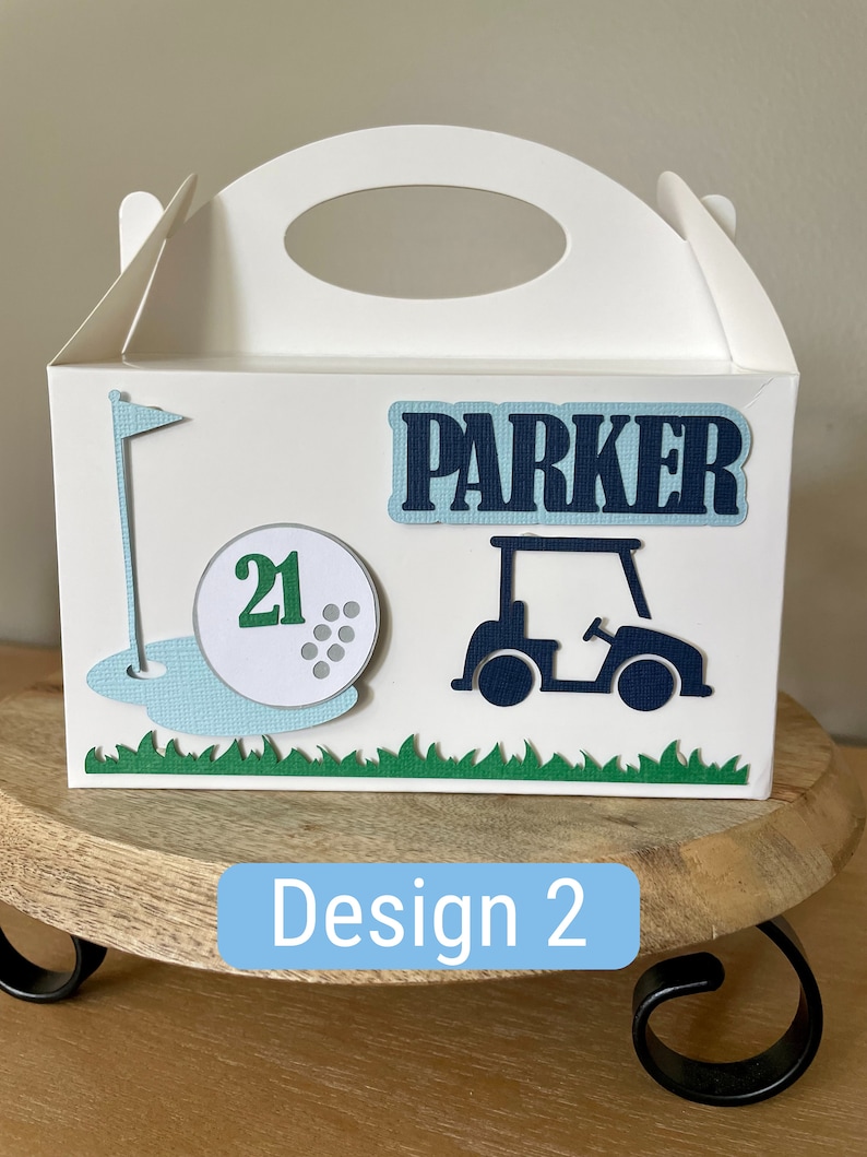 Puede incluir: Caja de regalo blanca con tem&aacute;tica de golf. La caja tiene una bola de golf verde y azul con el n&uacute;mero 21, una bandera de golf verde, un carrito de golf azul y el nombre "Parker" en la parte delantera.