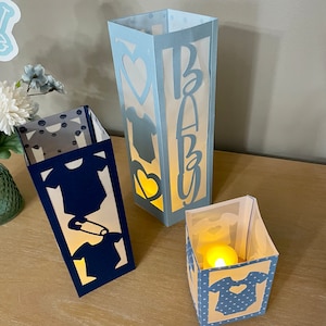 Op de afbeelding: Drie papieren lantaarns met blauwe en witte stippenpatronen en uitgesneden ontwerpen. De lantaarns zijn versierd met een baby-romper, een hart en het woord "Baby".