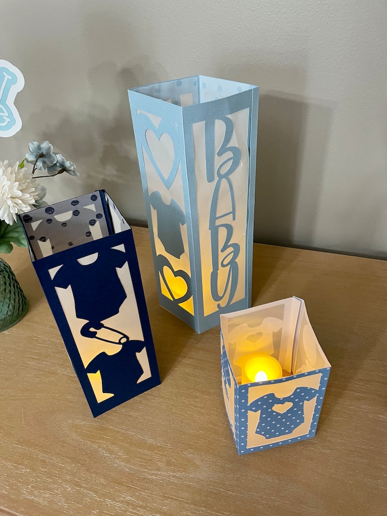 Op de afbeelding: Drie papieren lantaarns met blauwe en witte stippenpatronen en uitgesneden ontwerpen. De lantaarns zijn versierd met een baby-romper, een hart en het woord "Baby".