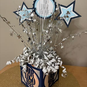 Puede incluir: Un centro de mesa azul marino y blanco con detalles de estrellas y círculos. El centro de mesa tiene el texto "YOUR LOGO OR TEXT HERE" y está decorado con confeti plateado en forma de estrella y serpentinas plateadas. La base es una caja azul marino.