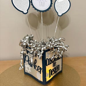 May include: A navy blue and silver themed centerpiece. It features a box with the text "Parker Customize the Design", topped with silver ribbon and three balloon decorations. The centerpiece is on a gold surface.