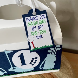 May include: White favor box with a blue and green golf-themed design. The box has a handle and a tag that says "Thanks for swinging by my par-tee! Love."