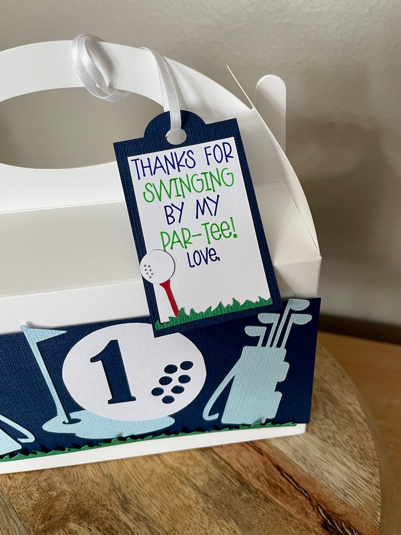 May include: White favor box with a blue and green golf-themed design. The box has a handle and a tag that says "Thanks for swinging by my par-tee! Love."