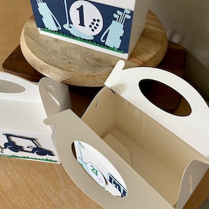 Puede incluir: Tres cajas de regalo blancas con dise&ntilde;os de tem&aacute;tica de golf. Las cajas tienen asas recortadas y est&aacute;n decoradas con pelotas de golf, palos de golf y carritos de golf. Las cajas est&aacute;n numeradas 1, 2 y 21.