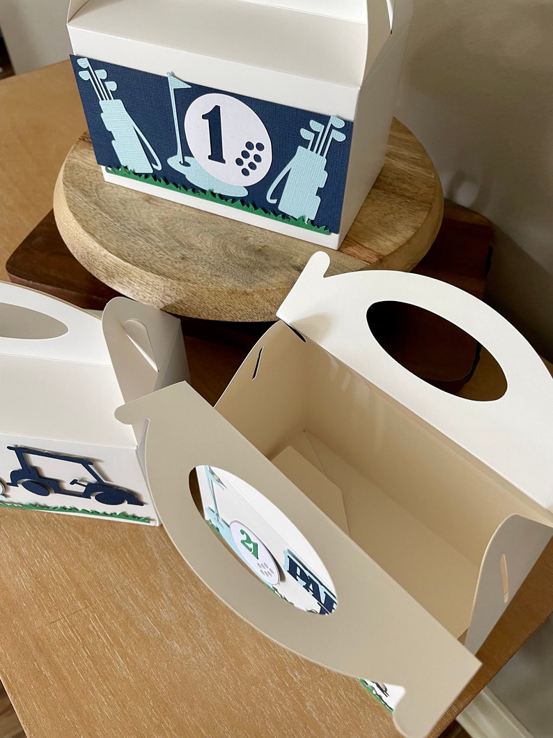 Puede incluir: Tres cajas de regalo blancas con dise&ntilde;os de tem&aacute;tica de golf. Las cajas tienen asas recortadas y est&aacute;n decoradas con pelotas de golf, palos de golf y carritos de golf. Las cajas est&aacute;n numeradas 1, 2 y 21.
