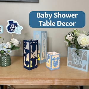 Op de afbeelding: Blauwe en witte papieren lantaarns met uitgesneden ontwerpen van babykleding, fopspeen en de woorden "Sweet Baby Boy". De lantaarns staan op een houten tafel met een boeket witte rozen en blauwe bloemen in een vaas.