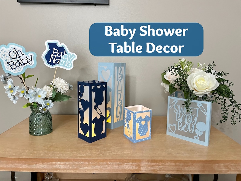 Op de afbeelding: Blauwe en witte papieren lantaarns met uitgesneden ontwerpen van babykleding, fopspeen en de woorden "Sweet Baby Boy". De lantaarns staan op een houten tafel met een boeket witte rozen en blauwe bloemen in een vaas.
