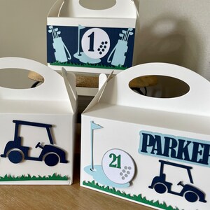 Puede incluir: Tres cajas de regalo blancas con decoraci&oacute;n de tem&aacute;tica de golf. Las cajas tienen asas recortadas y est&aacute;n decoradas con carritos de golf, pelotas de golf y palos de golf. Una caja tiene el n&uacute;mero "1", otra el n&uacute;mero "21" y la tercera el nombre "Parker".