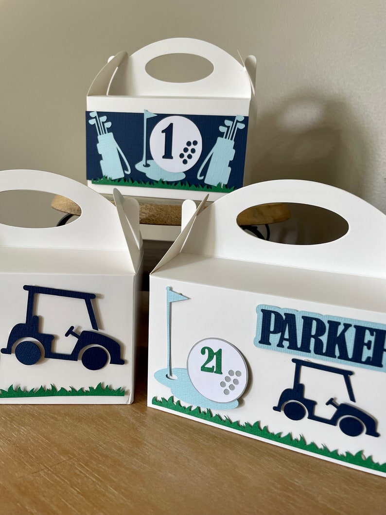 Puede incluir: Tres cajas de regalo blancas con decoraci&oacute;n de tem&aacute;tica de golf. Las cajas tienen asas recortadas y est&aacute;n decoradas con carritos de golf, pelotas de golf y palos de golf. Una caja tiene el n&uacute;mero "1", otra el n&uacute;mero "21" y la tercera el nombre "Parker".