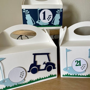 Puede incluir: Tres cajas de regalo blancas con dise&ntilde;os de tem&aacute;tica de golf. Las cajas tienen asas recortadas y est&aacute;n decoradas con pelotas de golf, palos de golf y carritos de golf. Las cajas est&aacute;n etiquetadas con los n&uacute;meros "1", "21" y "21".