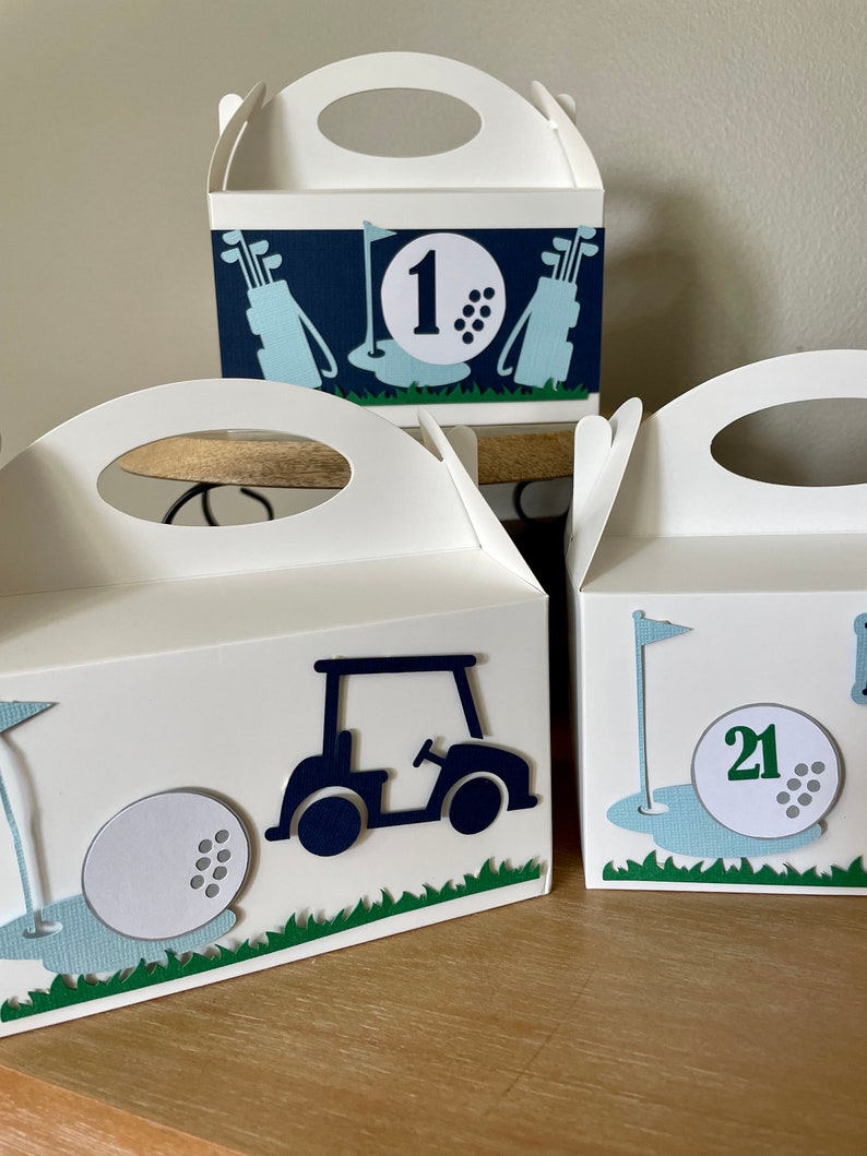 Puede incluir: Tres cajas de regalo blancas con dise&ntilde;os de tem&aacute;tica de golf. Las cajas tienen asas recortadas y est&aacute;n decoradas con pelotas de golf, palos de golf y carritos de golf. Las cajas est&aacute;n etiquetadas con los n&uacute;meros "1", "21" y "21".