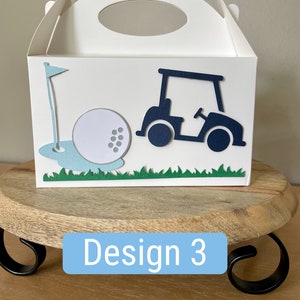 Puede incluir: Caja de regalo blanca con un dise&ntilde;o de carro de golf, pelota de golf y bandera. La caja tiene un asa azul y est&aacute; etiquetada como "Dise&ntilde;o 3".
