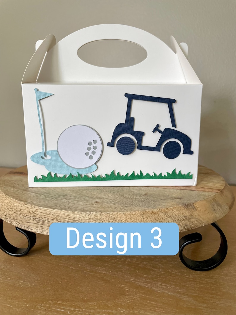 Puede incluir: Caja de regalo blanca con un dise&ntilde;o de carro de golf, pelota de golf y bandera. La caja tiene un asa azul y est&aacute; etiquetada como "Dise&ntilde;o 3".