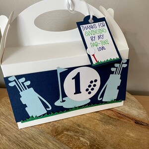 May include: White favor box with a blue and green golf-themed design. The box has a handle and a tag that says "Thanks for swinging by my par-tee! Love."