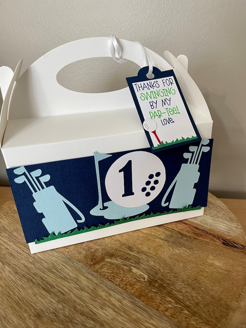 May include: White favor box with a blue and green golf-themed design. The box has a handle and a tag that says "Thanks for swinging by my par-tee! Love."