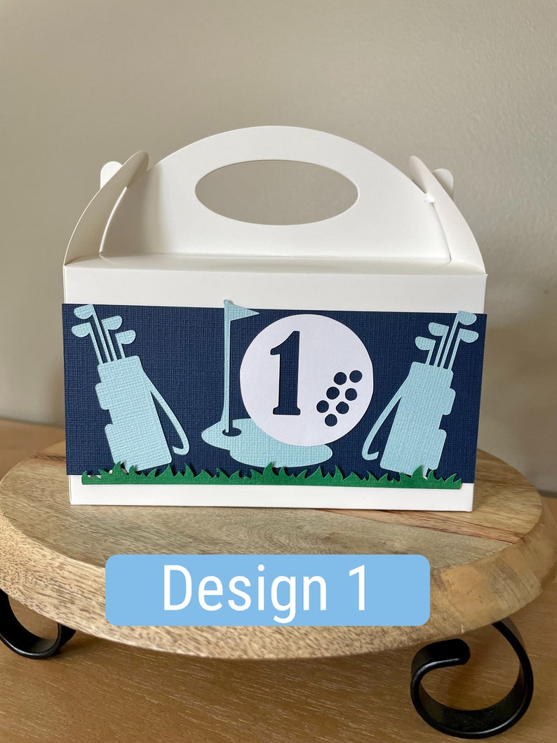 Puede incluir: Una caja de cart&oacute;n blanca con asa, decorada con un tema de golf. La caja tiene un fondo azul con un borde de hierba verde. El dise&ntilde;o presenta una bolsa de golf, palos de golf, una bola de golf con el n&uacute;mero "1" en ella y una bandera de golf.
