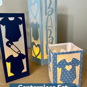 Op de afbeelding: Een set van drie blauwe en witte papieren lantaarns met een baby-rompertjesontwerp. De lantaarns zijn versierd met een hart en het woord "Baby".