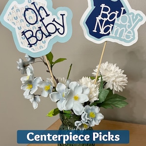 Op de afbeelding: Blauwe en witte baby shower-centerpieces met de tekst "Oh Baby" en "Baby Name". De picks zijn in een glazen vaas met witte bloemen.
