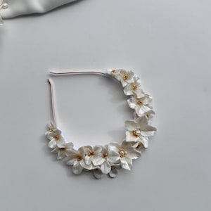 May include: A white floral headband with gold accents. The headband is made of fabric flowers and is perfect for a wedding or other special occasion.