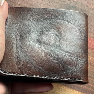 Puede incluir: Una cartera de cuero marrón con una superficie texturizada. La cartera es rectangular y parece estar doblada. Los bordes están cosidos con un hilo en contraste. La cartera está sujeta por una mano.