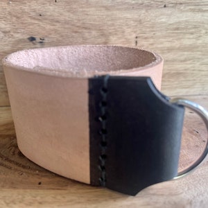 Puede incluir: Un portavasos de cuero hecho a mano con un cuerpo de color marrón claro natural y un asa negra. El asa está cosida al costado del portavasos y presenta un anillo de metal plateado. El portavasos está diseñado para sostener una taza de tamaño estándar.