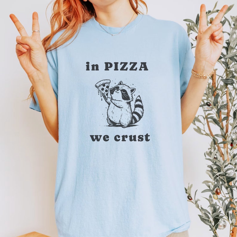 May include: A light blue t-shirt with a black and white illustration of a raccoon holding a slice of pizza. The text "in pizza we crust" is printed on the shirt.