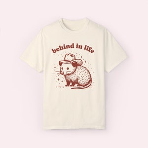 May include: A white t-shirt with a brown illustration of an opossum wearing a cowboy hat. The text "behind in life" is printed above the opossum.