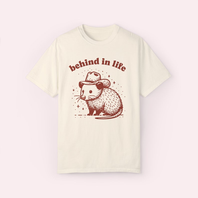May include: A white t-shirt with a brown illustration of an opossum wearing a cowboy hat. The text "behind in life" is printed above the opossum.