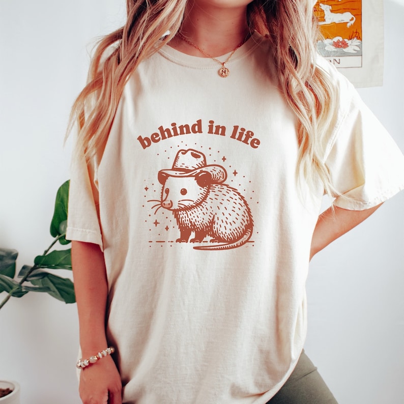 May include: A white t-shirt with a brown illustration of an opossum wearing a cowboy hat. The text "behind in life" is printed in brown above the opossum.