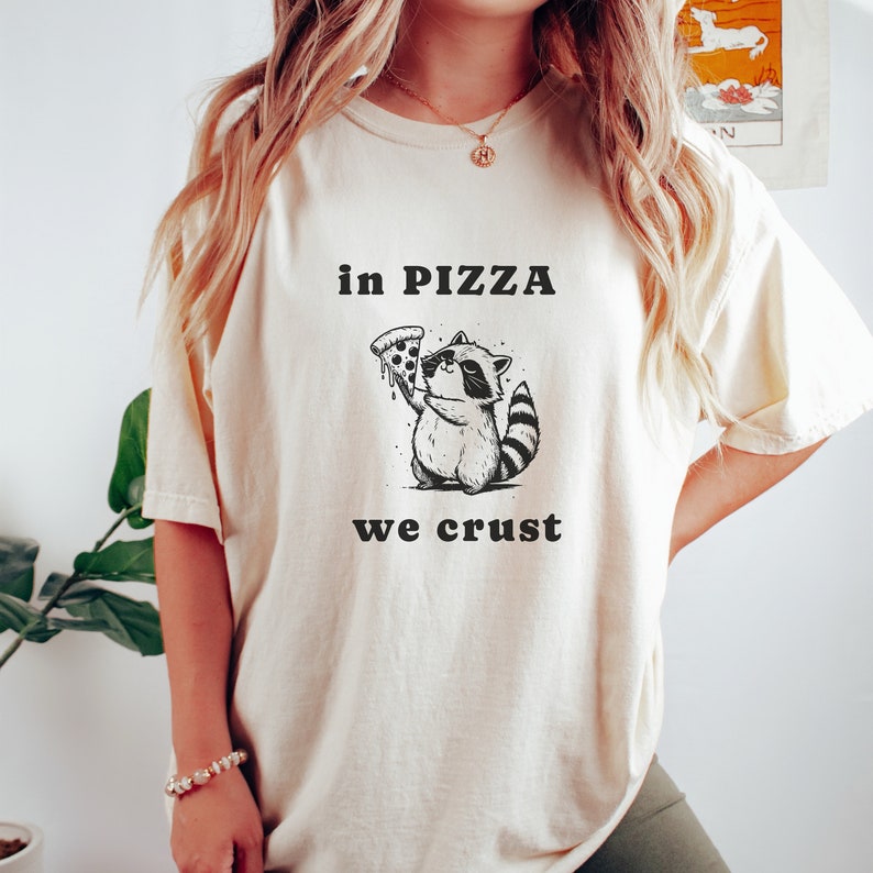 May include: A white t-shirt with a black graphic of a raccoon holding a pizza slice. The text "in pizza we crust" is printed below the raccoon.