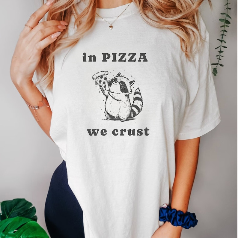 May include: White t-shirt with a black and white illustration of a raccoon holding a slice of pizza. The text "in pizza we crust" is printed below the illustration.