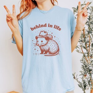 May include: A light blue t-shirt with a brown possum wearing a cowboy hat and the text "behind in life".