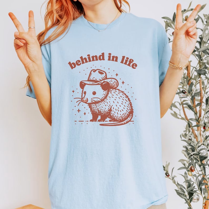 May include: A light blue t-shirt with a brown possum wearing a cowboy hat and the text "behind in life".