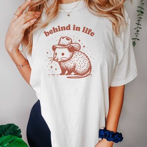 May include: White t-shirt with a brown illustration of an opossum wearing a cowboy hat. The text "behind in life" is printed above the opossum.