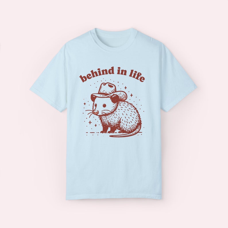 May include: Light blue t-shirt with a brown illustration of an opossum wearing a cowboy hat. The text "behind in life" is printed in brown above the opossum.
