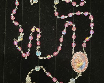 Könnte beinhalten: Ein Rosenkranz mit rosa irisierenden Perlen und einem silbernen Kruzifix. Der Rosenkranz hat eine silberne Kette und einen rosa ovalen Anhänger mit einem Bild von Maria und Jesus. Die Perlen sind durch eine silberne Kette verbunden.