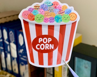 May include: A sticker of a red and white striped popcorn bucket filled with colorful planets with cute faces. The bucket says "POP CORN" in a white oval.