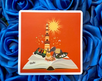 May include: An illustration of a red book with a yellow sun, a tower, an elephant, and a rickshaw. The book is open and the text "Delhi" is written on the page.