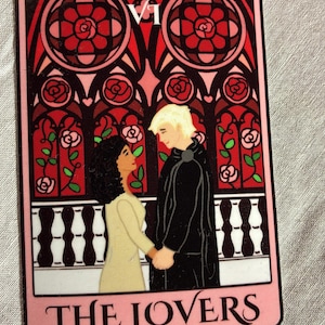 May include: A tarot card featuring a couple holding hands, set against a backdrop of red stained-glass windows with rose motifs. The card is framed in pink and has the words "THE LOVERS" at the bottom.