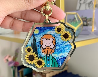 May include: A gold-toned keychain featuring a hexagonal acrylic charm with a cartoon illustration of Vincent van Gogh, the artist, with a blue and yellow background reminiscent of his famous Starry Night painting. The charm is surrounded by sunflowers.