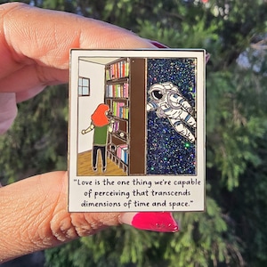 Puede incluir: Un pin de esmalte dorado con una ilustración colorida de una persona mirando una estantería y un astronauta flotando en el espacio. El pin incluye la cita "El amor es lo único que podemos percibir que trasciende las dimensiones del tiempo y el espacio."