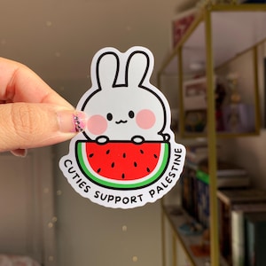 May include: A white bunny sticker with pink cheeks holding a red watermelon with black seeds. The sticker has the text "Cutie Support Palestine" around the watermelon.