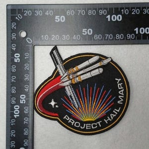 May include: Embroidered patch with a black background featuring a rocket design and the text "PROJECT HAIL MARY". The patch has a red and gold border, with blue, yellow, and red rays emanating from the bottom. A star is visible.