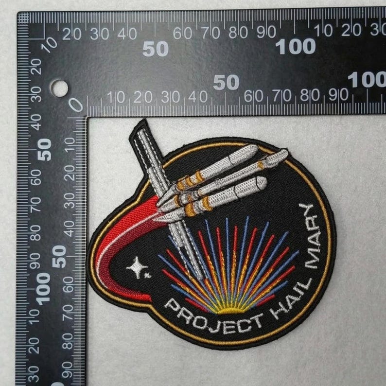 May include: Embroidered patch with a black background featuring a rocket design and the text "PROJECT HAIL MARY". The patch has a red and gold border, with blue, yellow, and red rays emanating from the bottom. A star is visible.