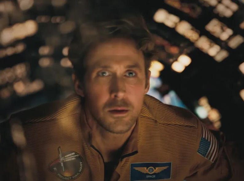 May include: A man wearing a tan space suit with patches, including an American flag and the word "SPACE". The man has a concerned expression, with a blurred background of lights.