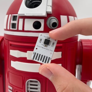 May include: A small white circuit board labelled "Droid Personality Chip" is held in front of a red and white robot. The chip has several small components and the text "Droid Personality Chip" printed on it. The robot has a cylindrical body.
